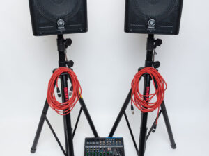 Zwei schwarze Yamaha Lautsprecher auf Stativen mit roten Kabeln, darunter ein Mischpult und mehrere Kabel und Adapter auf weißem Untergrund.