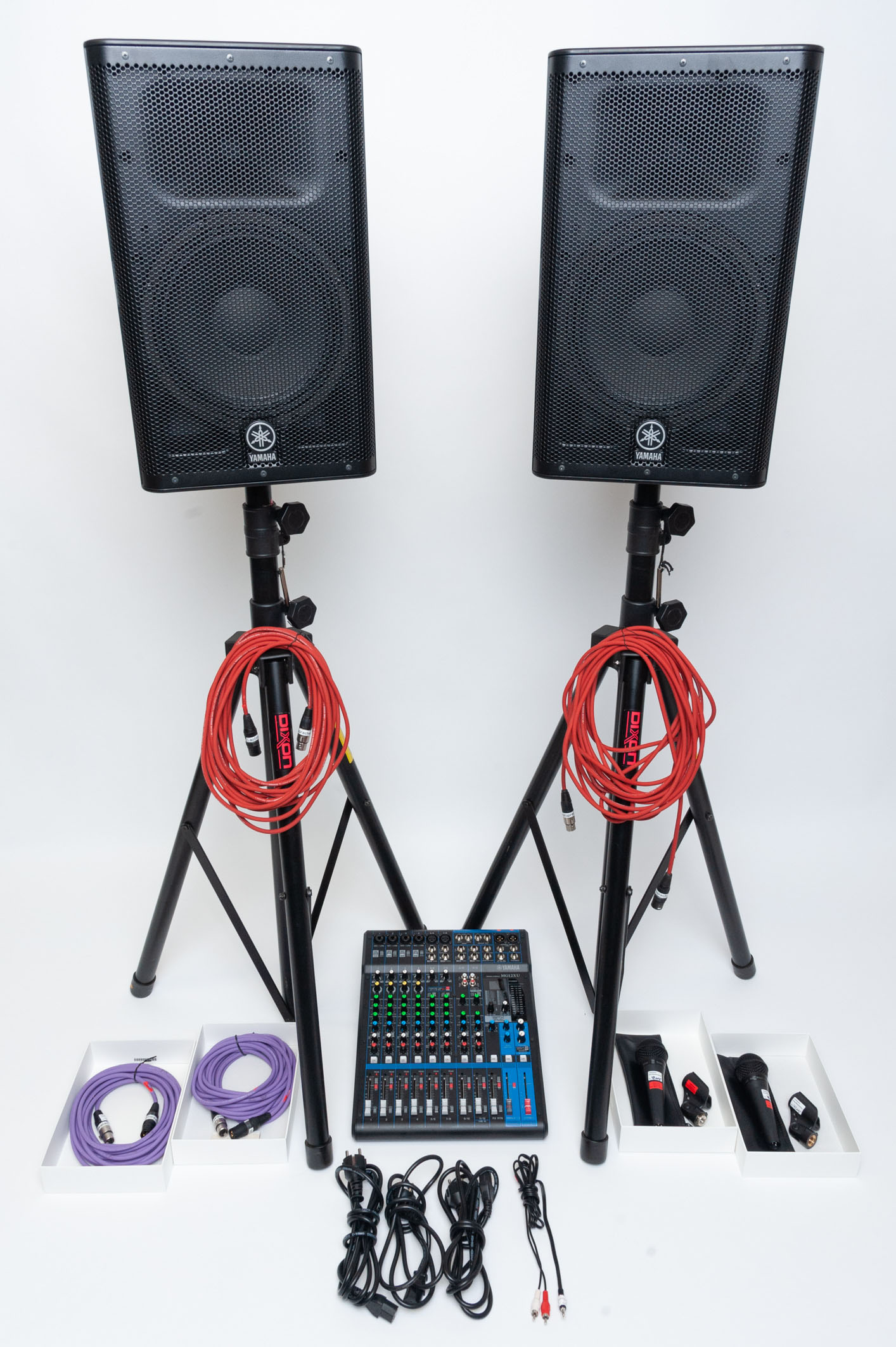 Zwei schwarze Yamaha Lautsprecher auf Stativen mit roten Kabeln, darunter ein Mischpult und mehrere Kabel und Adapter auf weißem Untergrund.