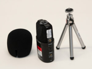 Schwarzes Zoom H2n Audiorecorder-Gerät mit Schaumstoff-Windschutz und kleinem silbernem Dreibein-Stativ auf hellem Untergrund.