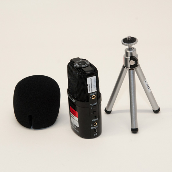 Schwarzes Zoom H2n Audiorecorder-Gerät mit Schaumstoff-Windschutz und kleinem silbernem Dreibein-Stativ auf hellem Untergrund.