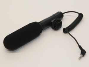 Schwarzes Richtmikrofon mit Schaumstoff-Windschutz und Spiralkabel mit Klinkenstecker auf hellem Untergrund.