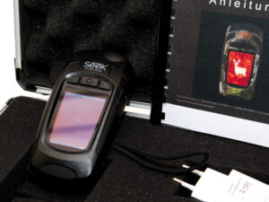 Schwarze Seek Wärmebildkamera mit Display, Bedienknöpfen und Trageschlaufe in schwarzem Schaumstoffkoffer, daneben Anleitung mit rotem Hirschbild und weißes Ladegerät.