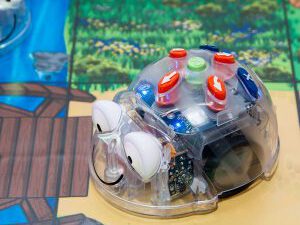 Transparenter Blue-Bot Lernroboter mit großen Augen auf einem Spielbrett mit Fluss und Holzbrücke.