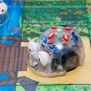 Transparenter Blue-Bot Lernroboter mit großen Augen auf einem Spielbrett mit Fluss und Holzbrücke.