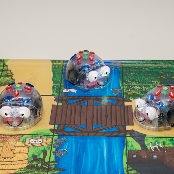 Drei transparente, Blue-Bot Lernroboter mit großen Augen auf einem Spielbrett mit Fluss und Holzbrücke.