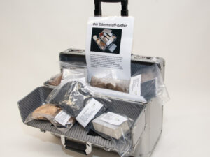 Offener silberner Aluminiumkoffer mit schwarzem Griff, darin mehrere durchsichtige Plastiktüten mit verschiedenen Dämmstoffproben und ein Informationsblatt mit dem Titel 'Der Dämmstoff-Koffer'
