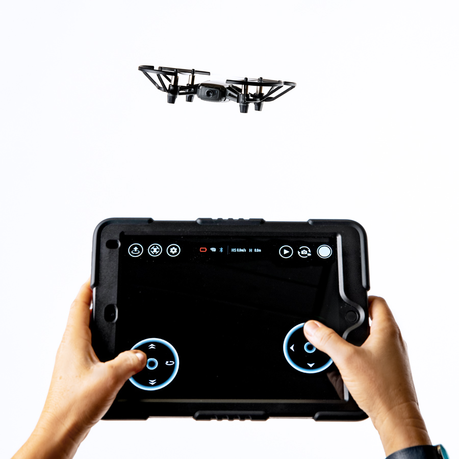 Zwei Hände steuern eine kleine schwarze Drohne mit einem Tablet, das Steuerkreise und Symbole zeigt.