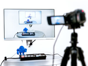 Kamera auf Stativ filmt einen kleinen blauen Roboter auf einem Tisch vor einem Bildschirm, der das Kamerabild zeigt.
