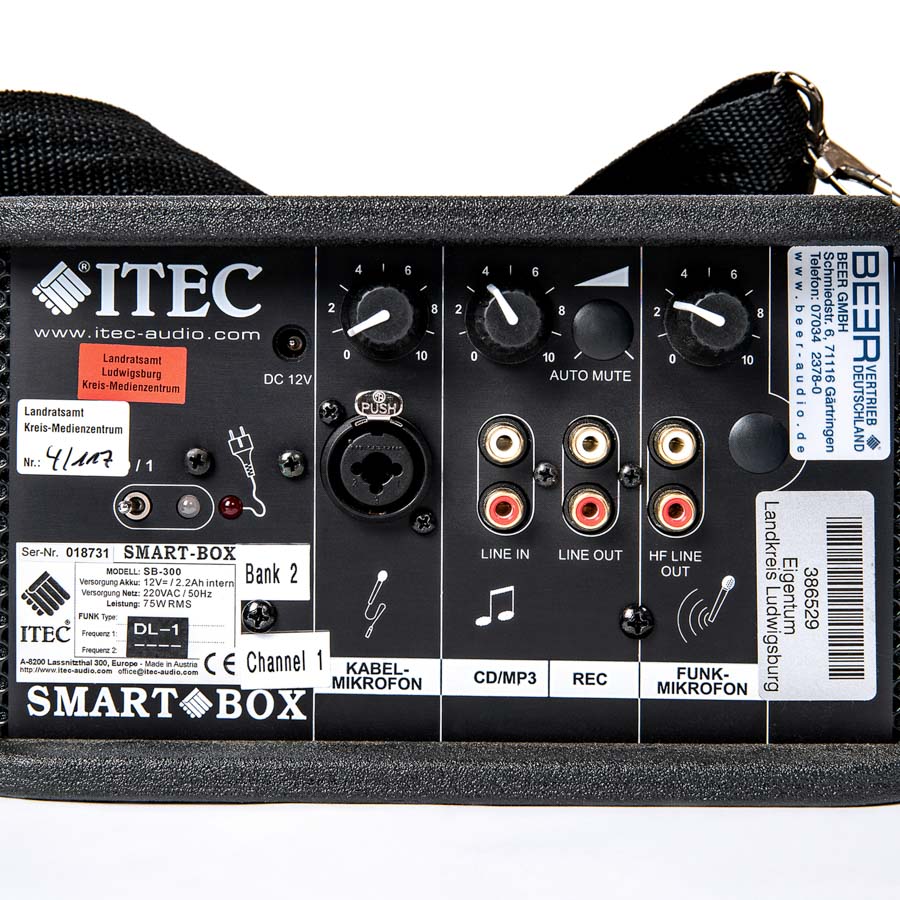Schwarzes ITEC SMART-BOX Audiogerät mit Drehreglern, Anschlüssen für Kabelmikrofon, CD/MP3, Aufnahme und Funkmikrofon, Aufklebern mit Texten und Barcodes.