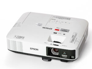 Ein weißer Epson-Beamer mit schwarzer Frontblende, auf der sich das Objektiv und ein HDMI-Logo befinden. Auf der Oberseite sind verschiedene Aufkleber und Beschriftungen sichtbar, darunter das Epson-Logo und Hinweise zur Bedienung. Der Beamer hat Lüftungsschlitze an der Seite und eine leicht abgerundete rechteckige Form.