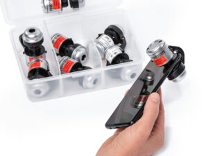 Hand hält Smartphone mit aufgesetztem Handymikroskop, daneben durchsichtige Box mit mehreren Mikroskopen mit roten Etiketten.