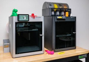 Zwei 3D-Drucker auf einem Holztisch, links ein silberner Bambu Lab X1 Carbon mit grünem und rotem 3D-gedrucktem Objekt oben, rechts ein schwarzer AMS P1S mit transparentem Filamentspender und mehreren Filamentrollen.