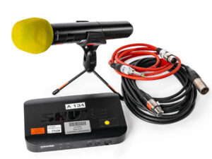 Drahtloses Shure SM58 Mikrofon mit gelbem Windschutz auf kleinem Stativ, daneben Shure Funksystem-Empfänger und zwei XLR-Kabel in Rot und Schwarz