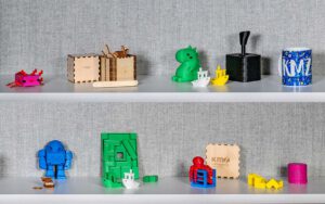 Mehrere bunte 3D-gedruckte Objekte in verschiedenen Formen und Größen auf einem weißen Regal.