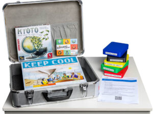 Offener silberner Klima-Spiele Koffer mit den Spielen "Kyoto", "Klimaxo" und "Keep Cool" auf einem weißen Tisch, daneben Stapel bunter Spielkarten und ein Blatt mit Text und QR-Code.