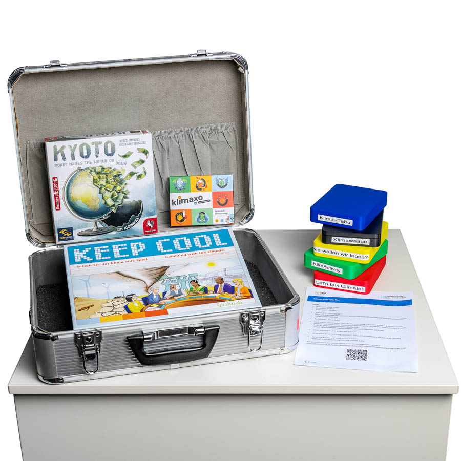 Offener silberner Klima-Spiele Koffer mit den Spielen "Kyoto", "Klimaxo" und "Keep Cool" auf einem weißen Tisch, daneben Stapel bunter Spielkarten und ein Blatt mit Text und QR-Code.