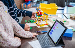 Ansicht eines Laptops und eines erstellten Lego Robotik Objekts an dem 2 Kinderhände arbeiten